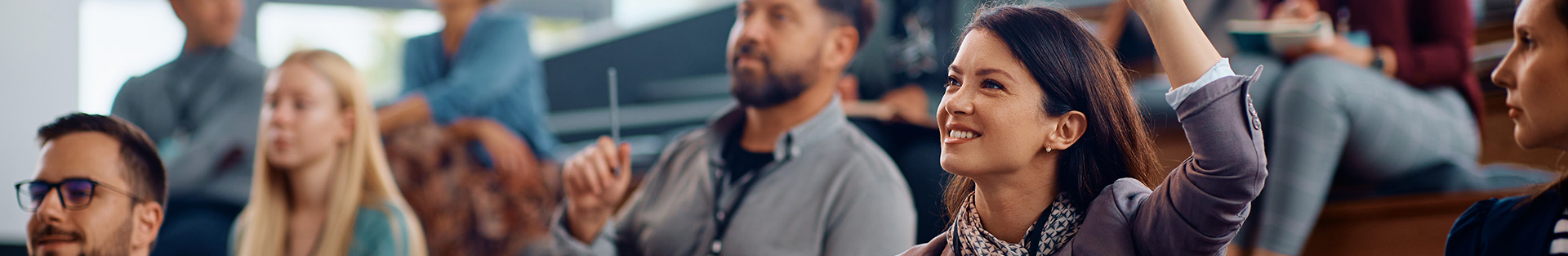 Grupa młodych ludzi siedzących w auli na spotkaniu i podnoszacych ręce w celu zadania pytania